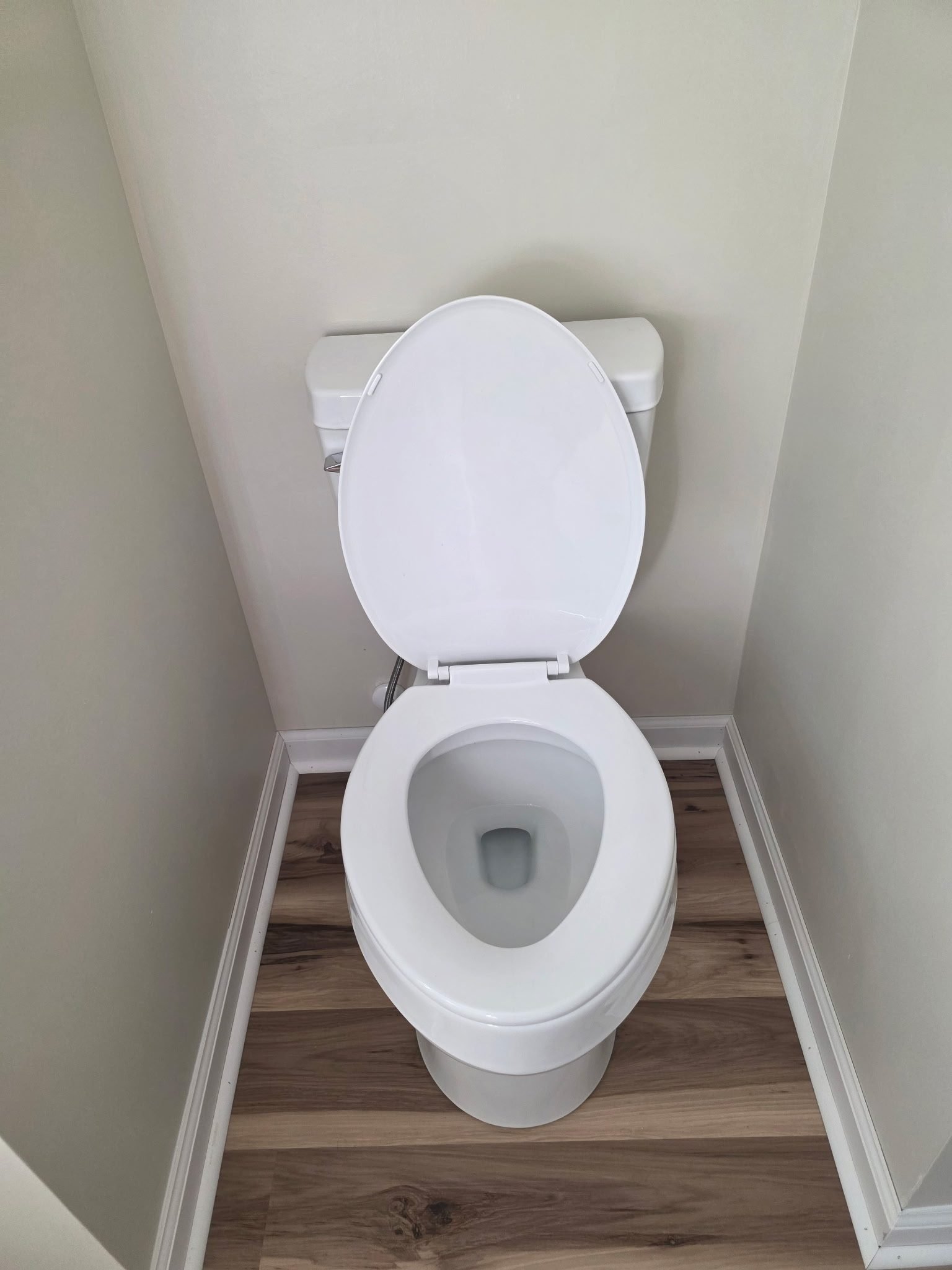 White elongated toilet, freshly installed in a corner bathroom with wood-look flooring.