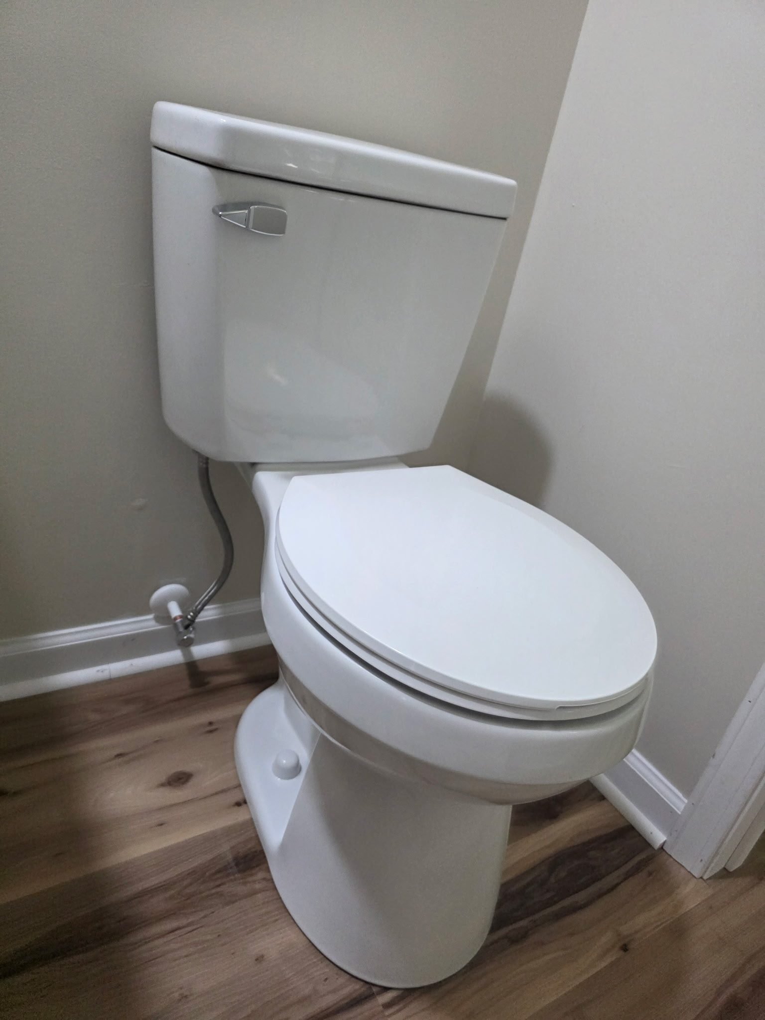 White toilet with chrome flush handle and supply line installed in the corner of a bathroom.
