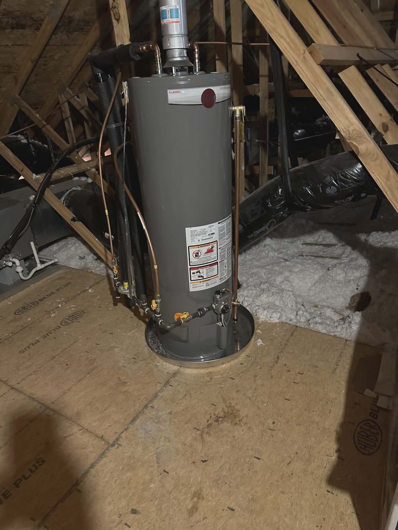 Water heater installed in an attic with copper piping and insulation among exposed roof framing.