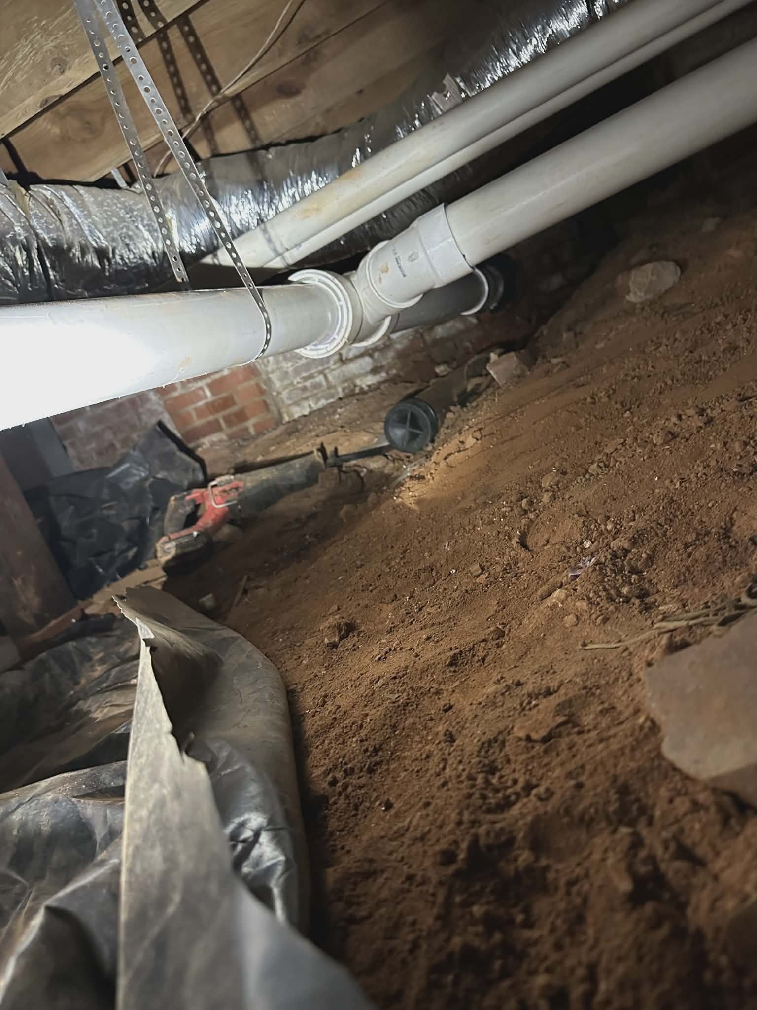 Crawlspace view of white PVC pipes running along floor joists above a dirt floor.