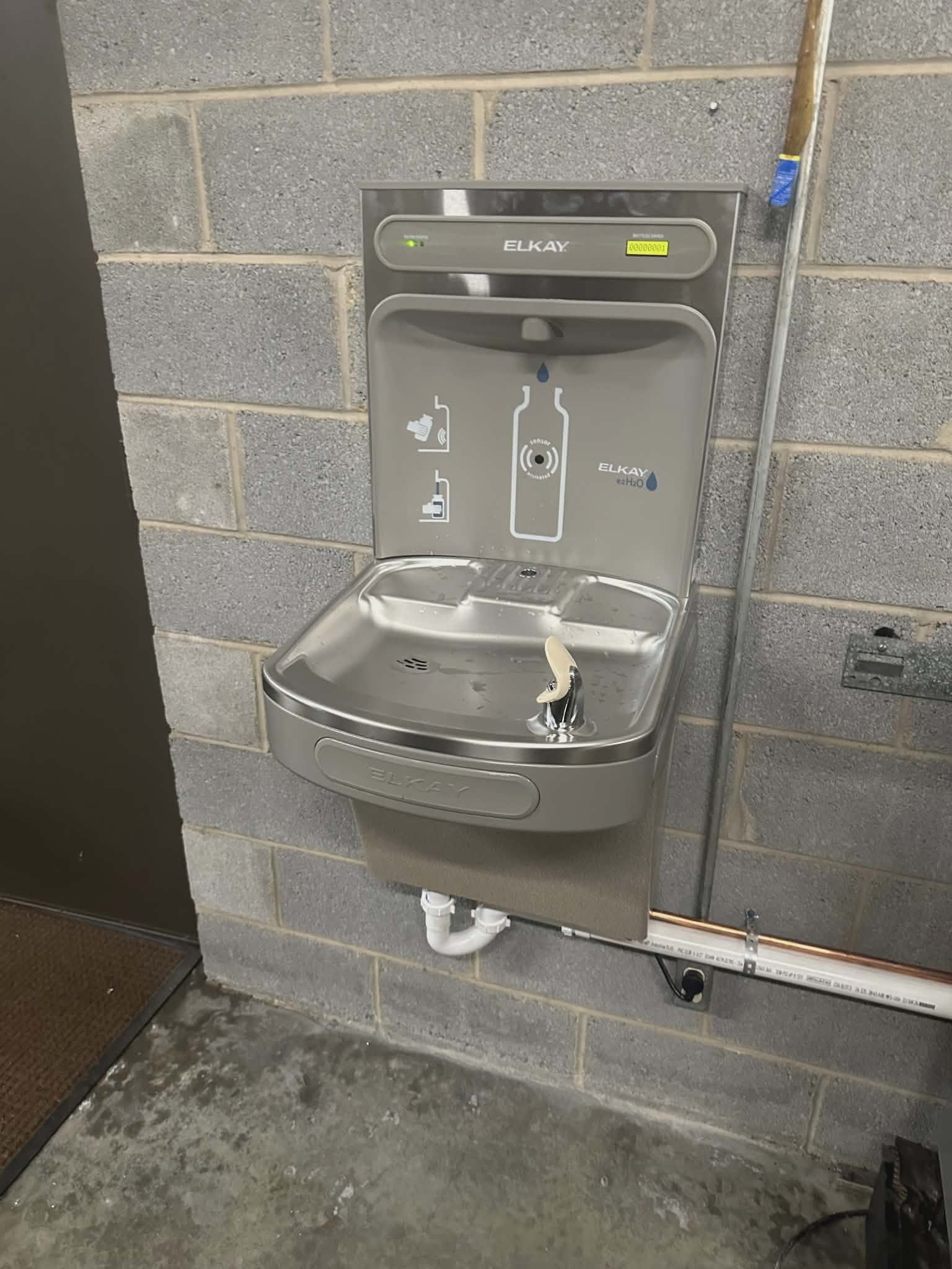 Stainless steel bottle-filling drinking fountain mounted on a wall with PVC drain piping below.