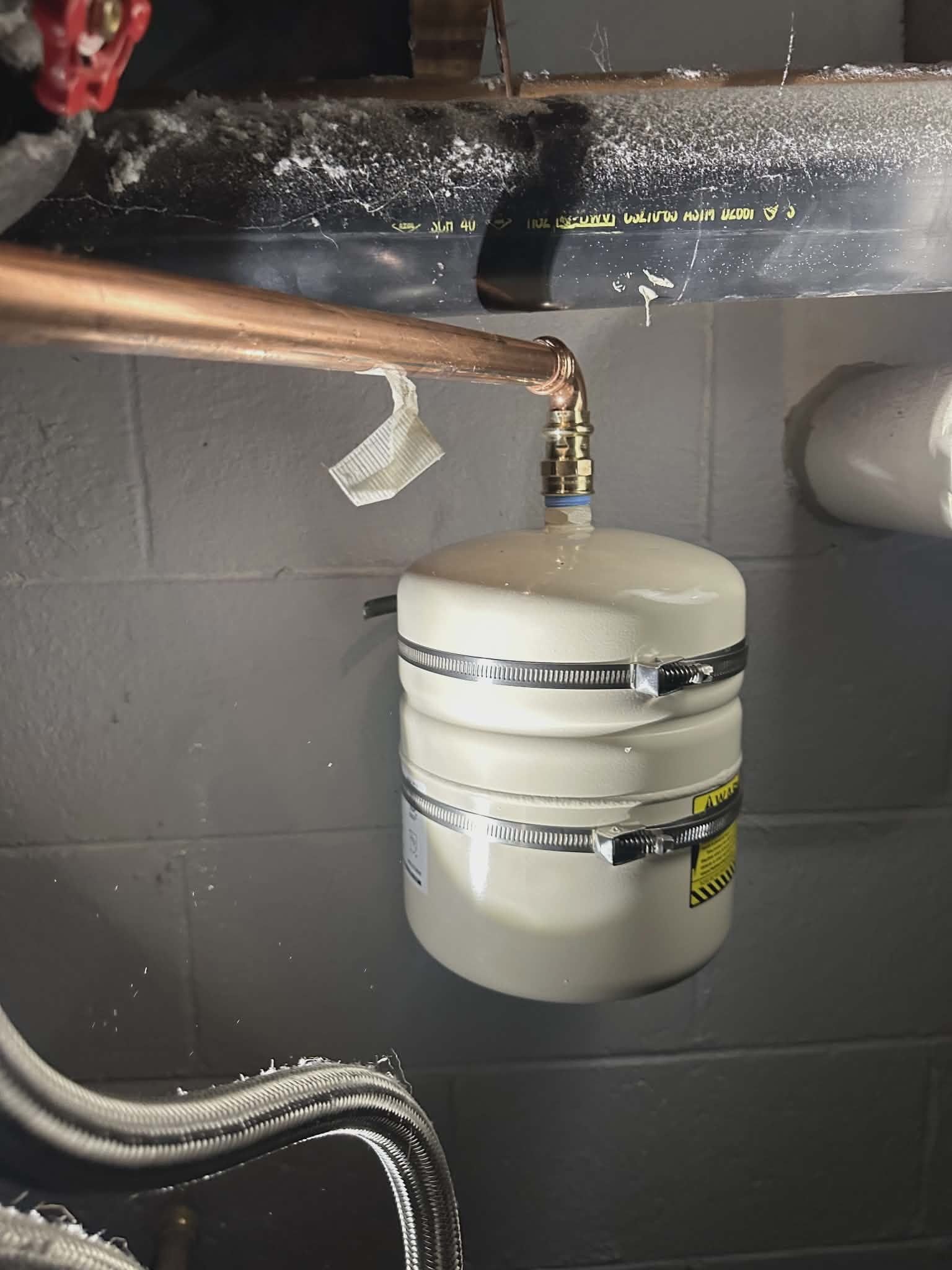 Small expansion tank with hose clamps mounted on copper piping against a wall.