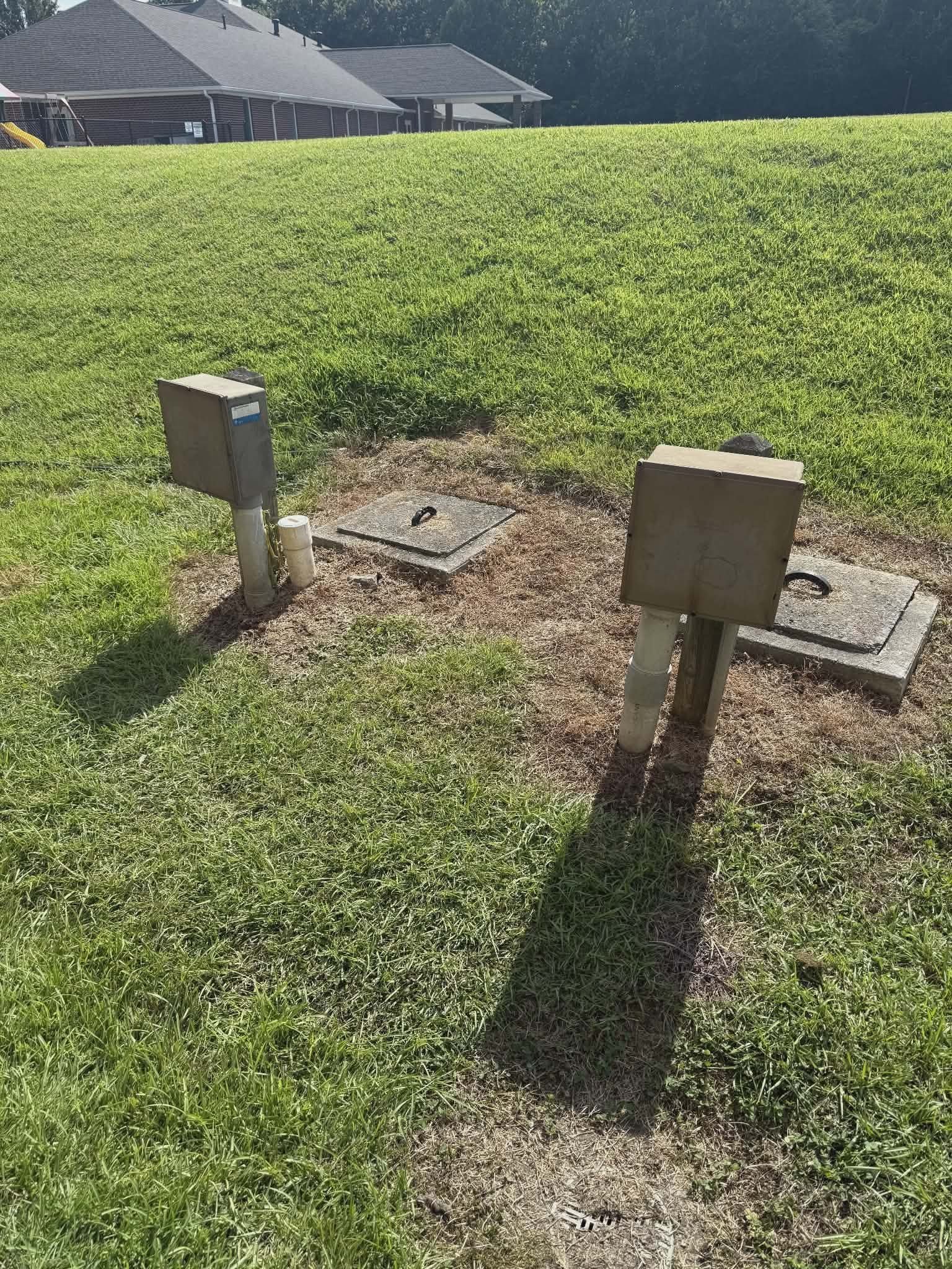 Two septic or well utility access boxes with PVC risers set in a lawn near a house.