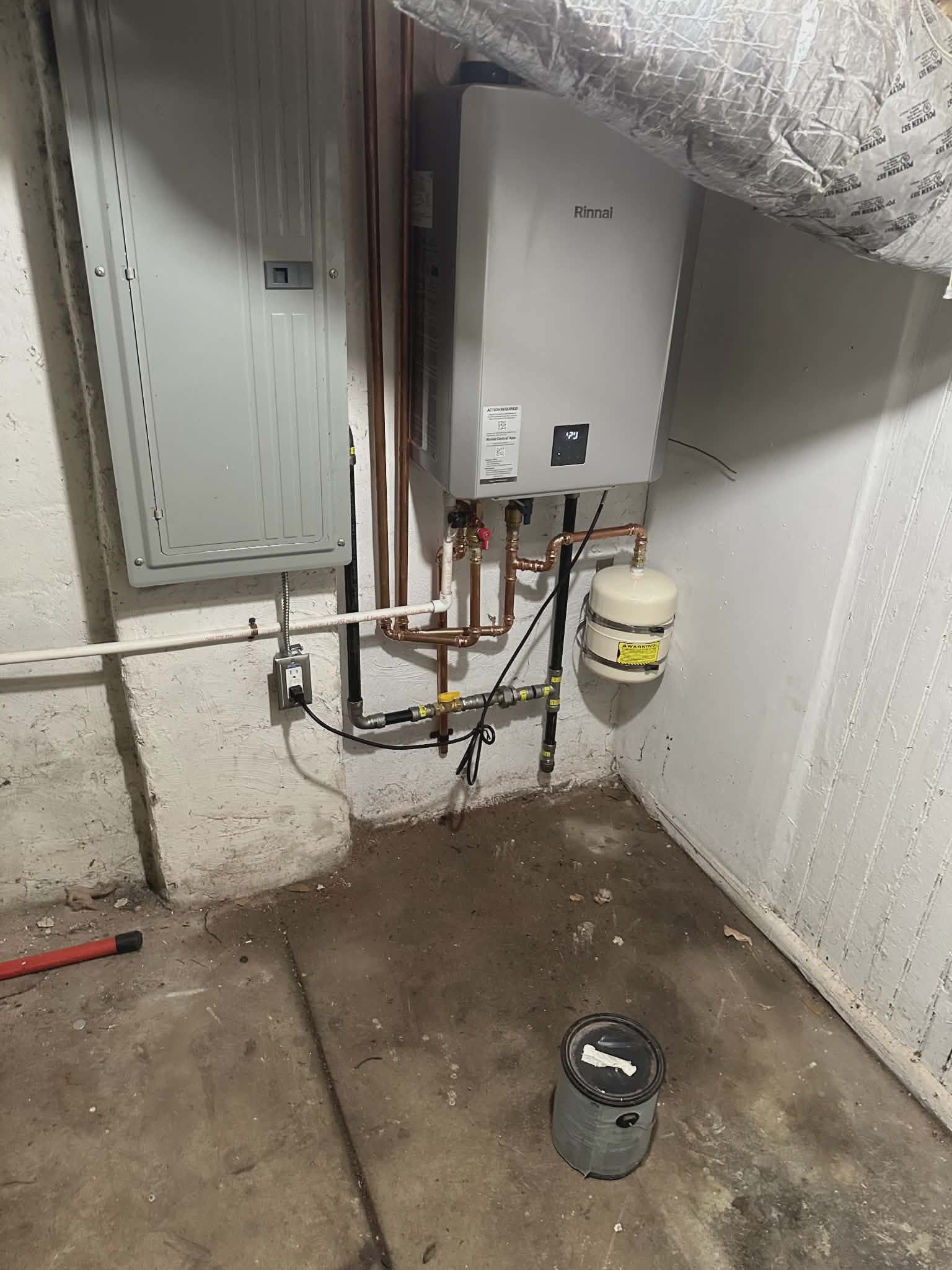 Tankless water heater mounted above an electrical panel with copper piping and a small expansion tank.