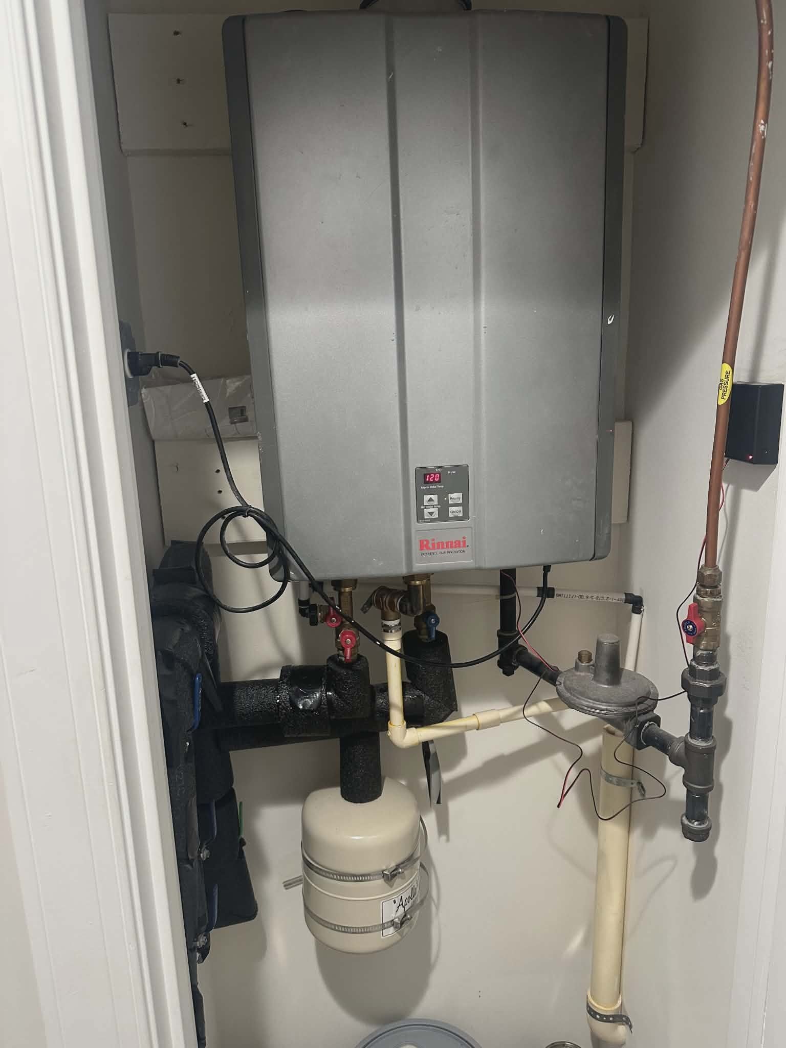 Tankless water heater installed in a closet with a small expansion tank and gas regulator below.