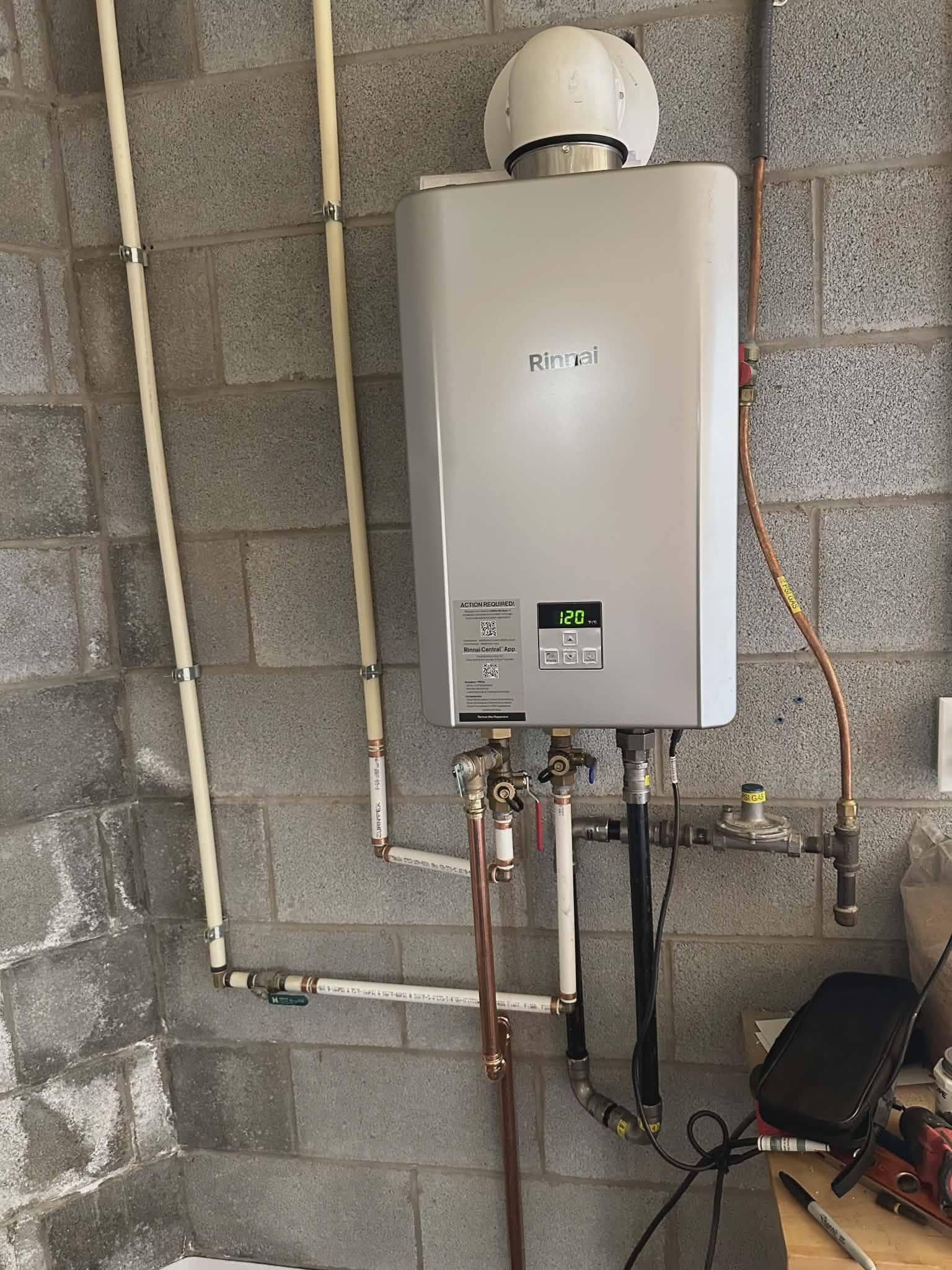 Tankless water heater mounted on a wall with copper and PVC supply piping and a gas regulator below.