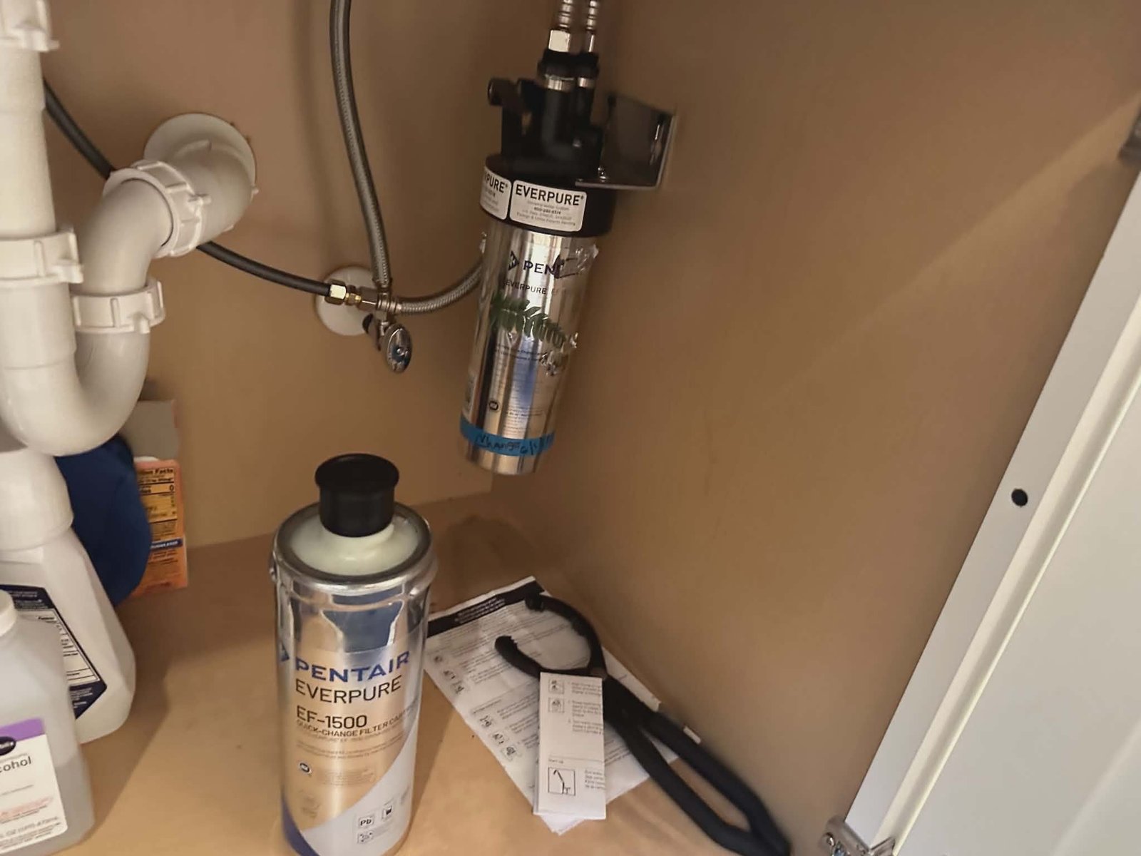 Inline water filter cartridge mounted under a kitchen sink next to white PVC drain piping.