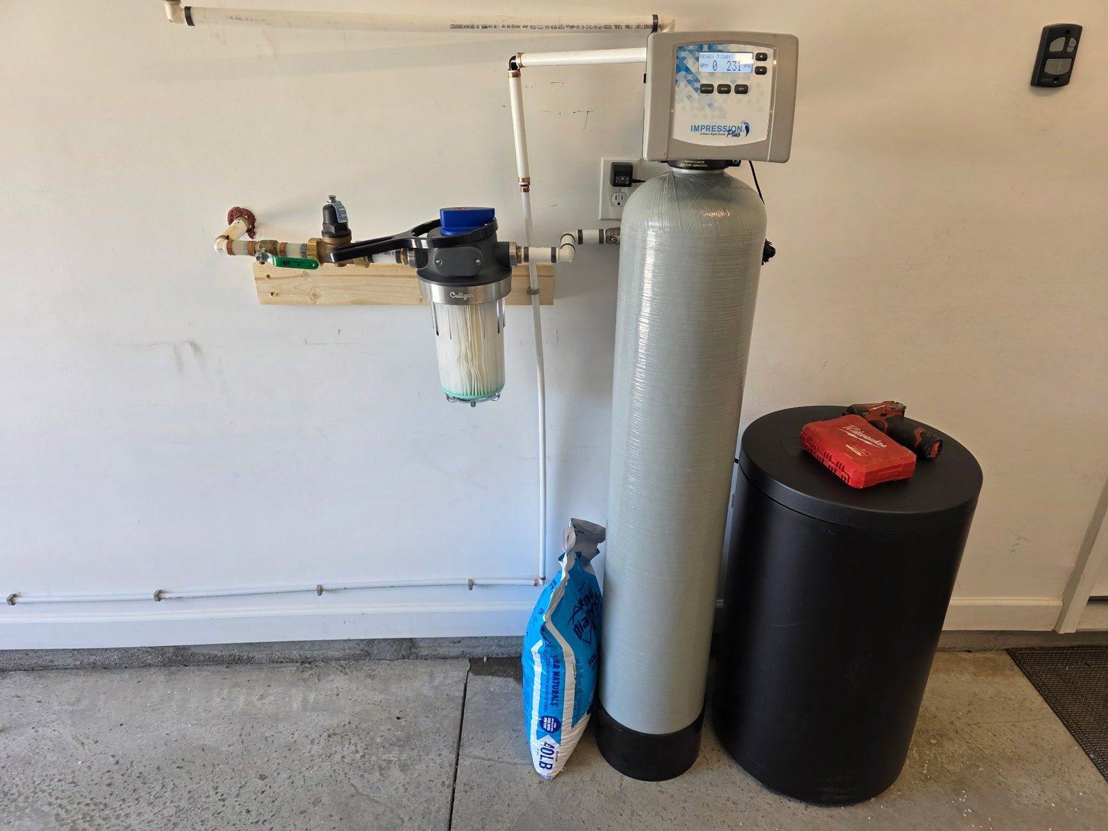 Water softener system with a brine tank, resin tank, digital control head, and whole-house filter mounted on a garage wall.