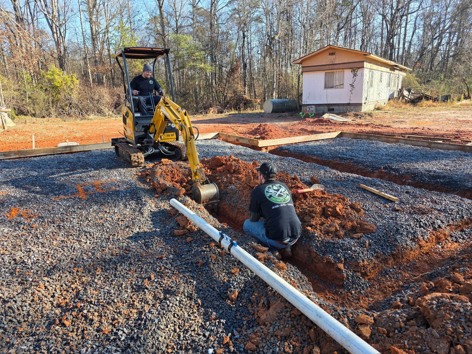Mini excavator digging a trench while a worker kneels beside a PVC pipe laid along a gravel building pad.
