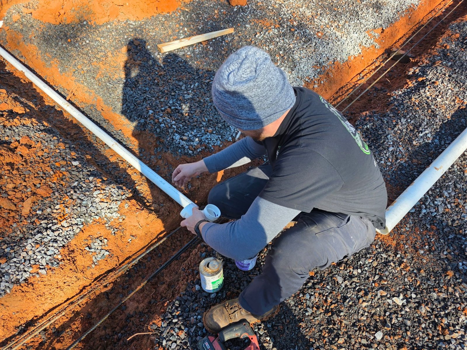 Overhead view of a worker kneeling on gravel gluing a fitting onto white PVC pipe at a trench junction.