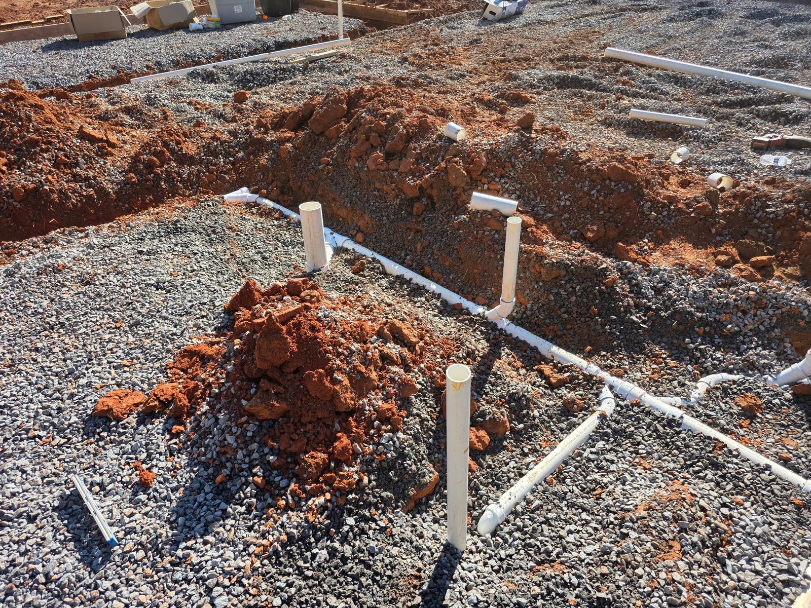 Overhead view of white PVC drain pipes with vertical stub-outs running through gravel-filled trenches.