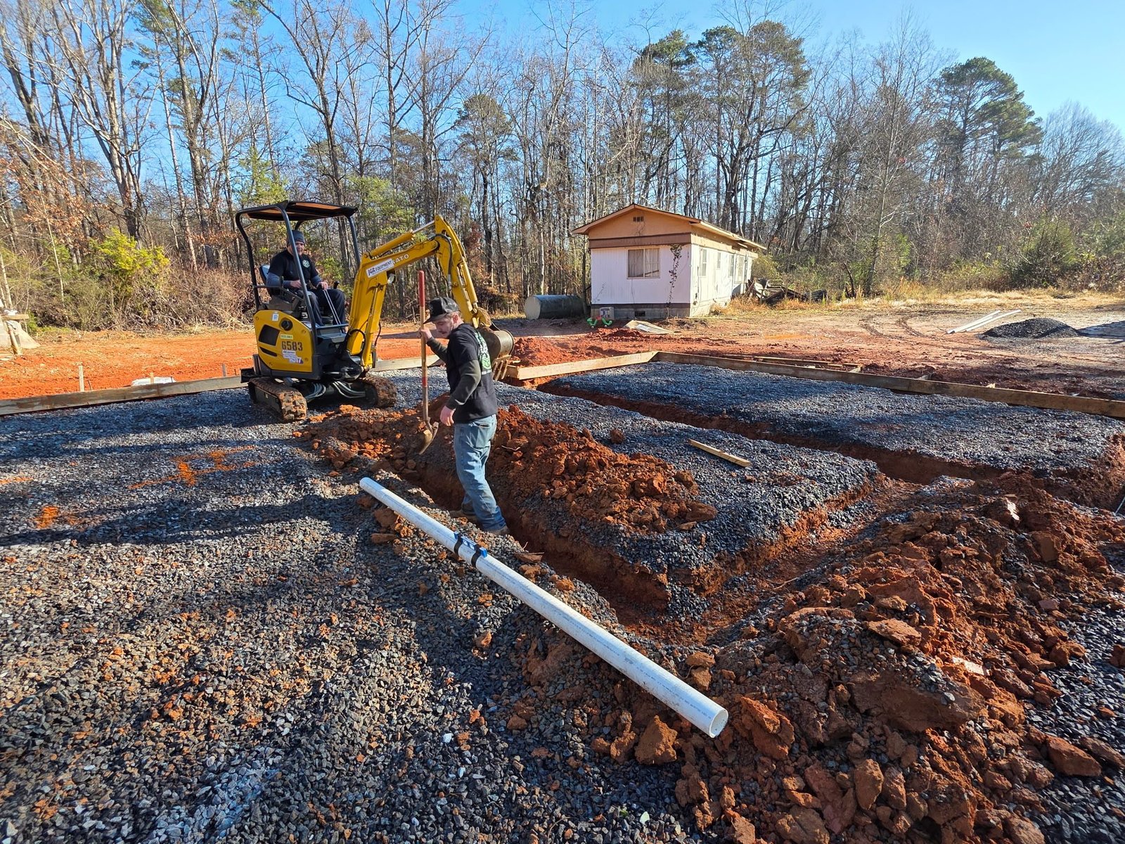 Worker standing in a freshly dug trench beside a mini excavator with PVC pipe laid along the gravel pad.