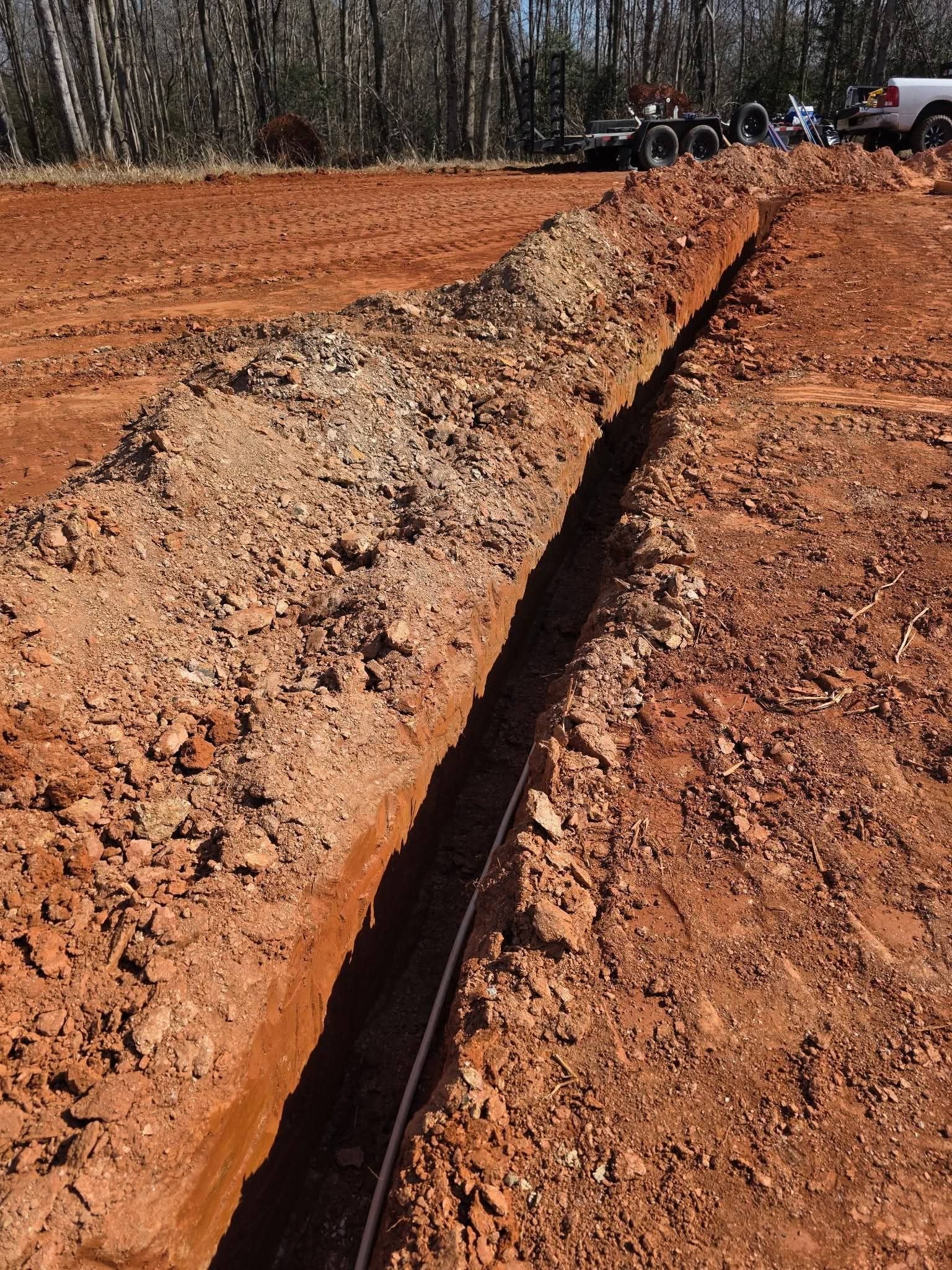 Deep trench with pipe at the bottom and equipment parked at the far end of the trench line.