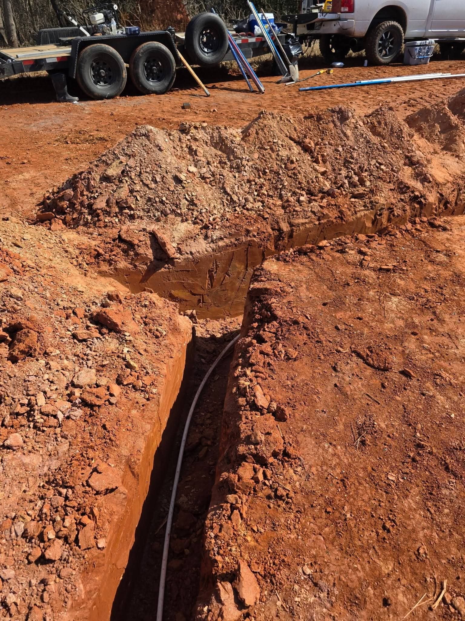 Close-up of a narrow trench with a pipe laid in the bottom and equipment staged above grade.