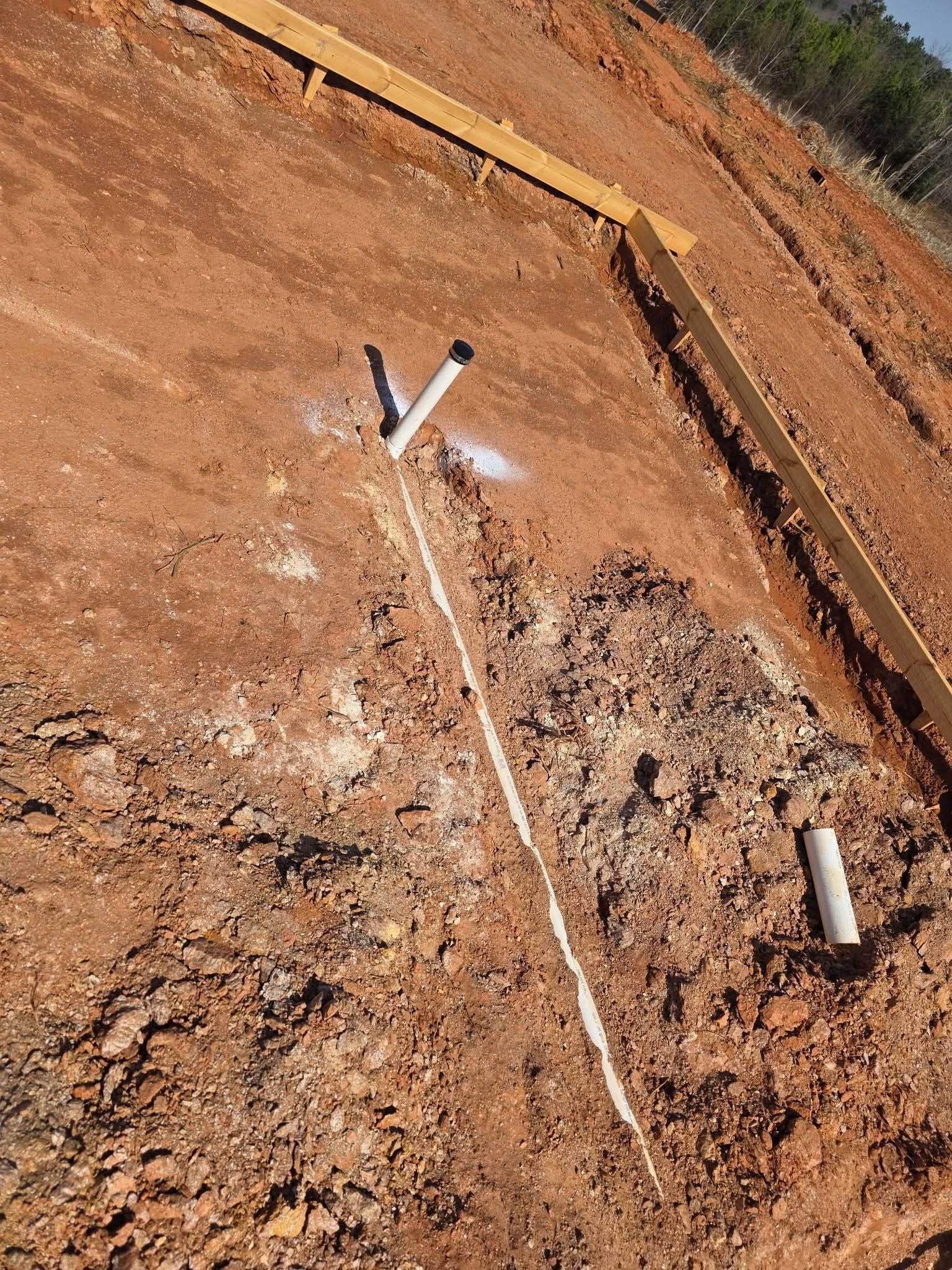 Two PVC pipe stubs and a connecting line partly backfilled with dirt next to a foundation form board.