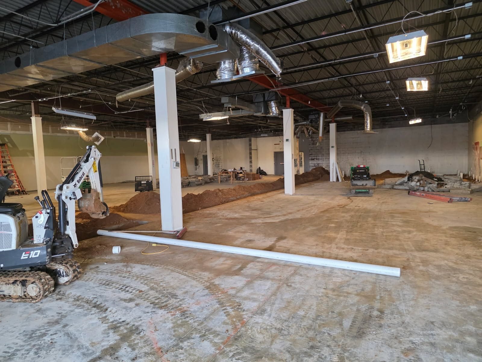 Open commercial building space with a mini excavator parked beside mounded dirt trenches.