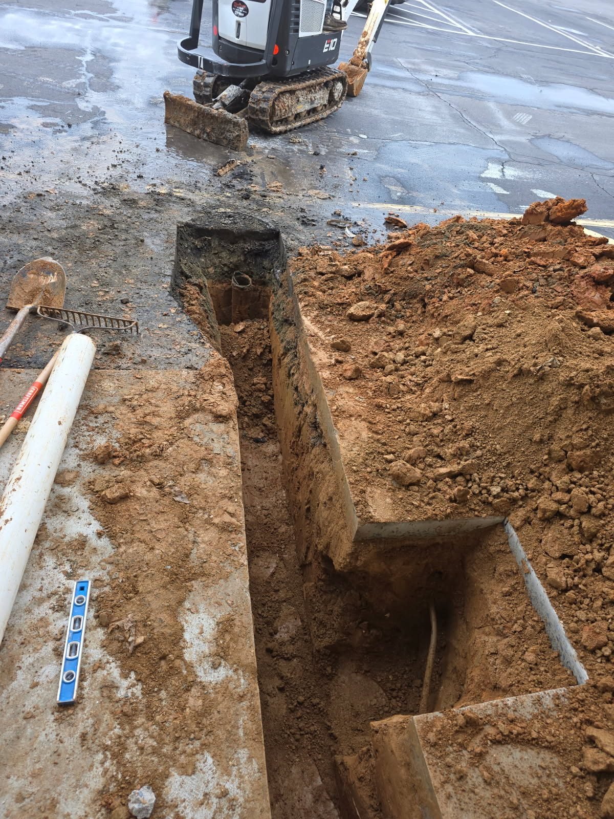 Open exterior trench with a mini excavator nearby and PVC pipe sections staged beside the dig on a commercial lot.