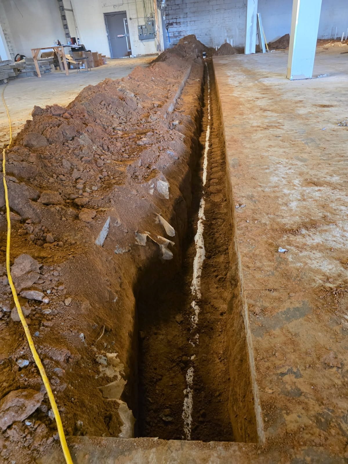 Long interior trench cut into the dirt subgrade of a commercial building with a PVC pipe run laid along the bottom.
