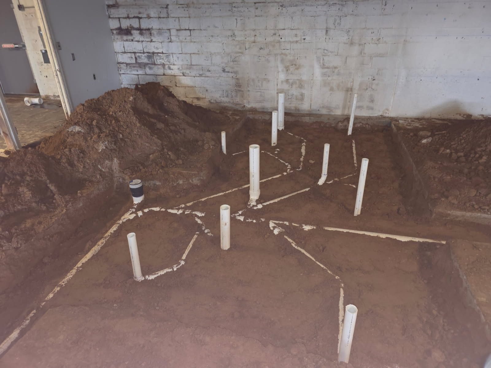 PVC stub-up pipes set across an excavated dirt floor near a doorway, with connecting pipe-run lines marked in the soil.