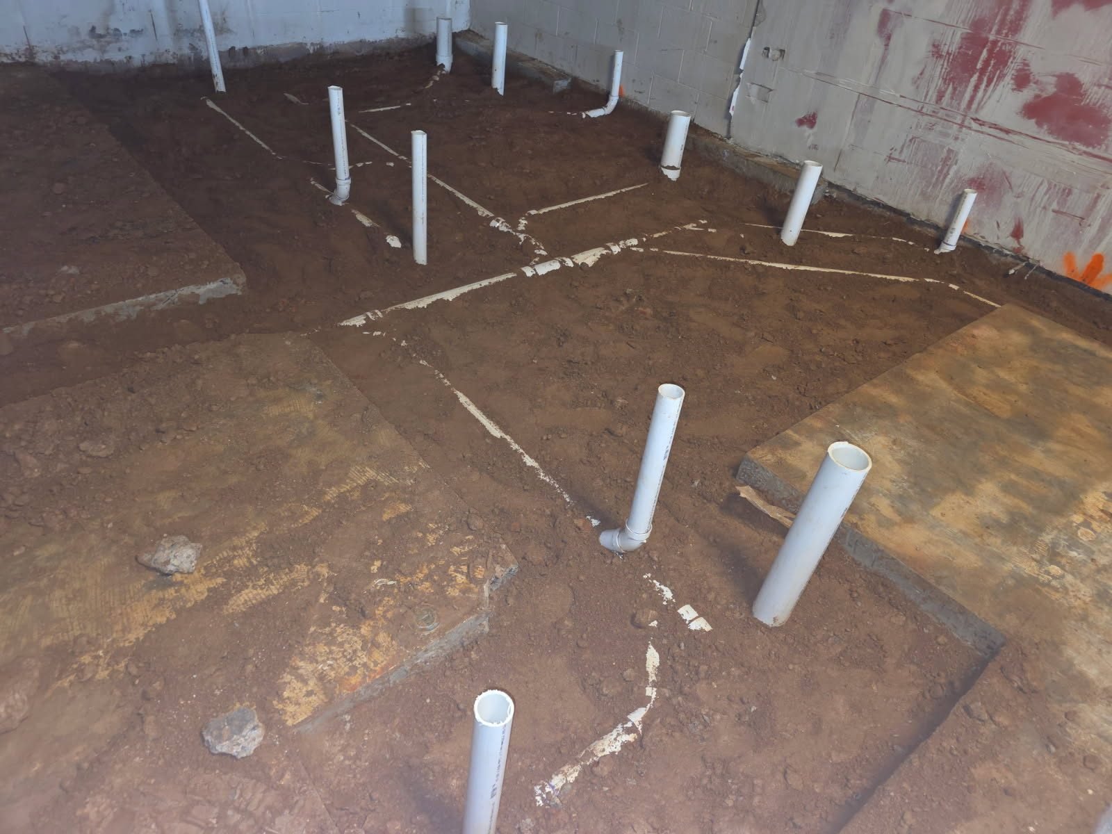 Overhead view of PVC stub-up pipes set in dirt with chalked layout lines marking the pipe network before the slab pour.