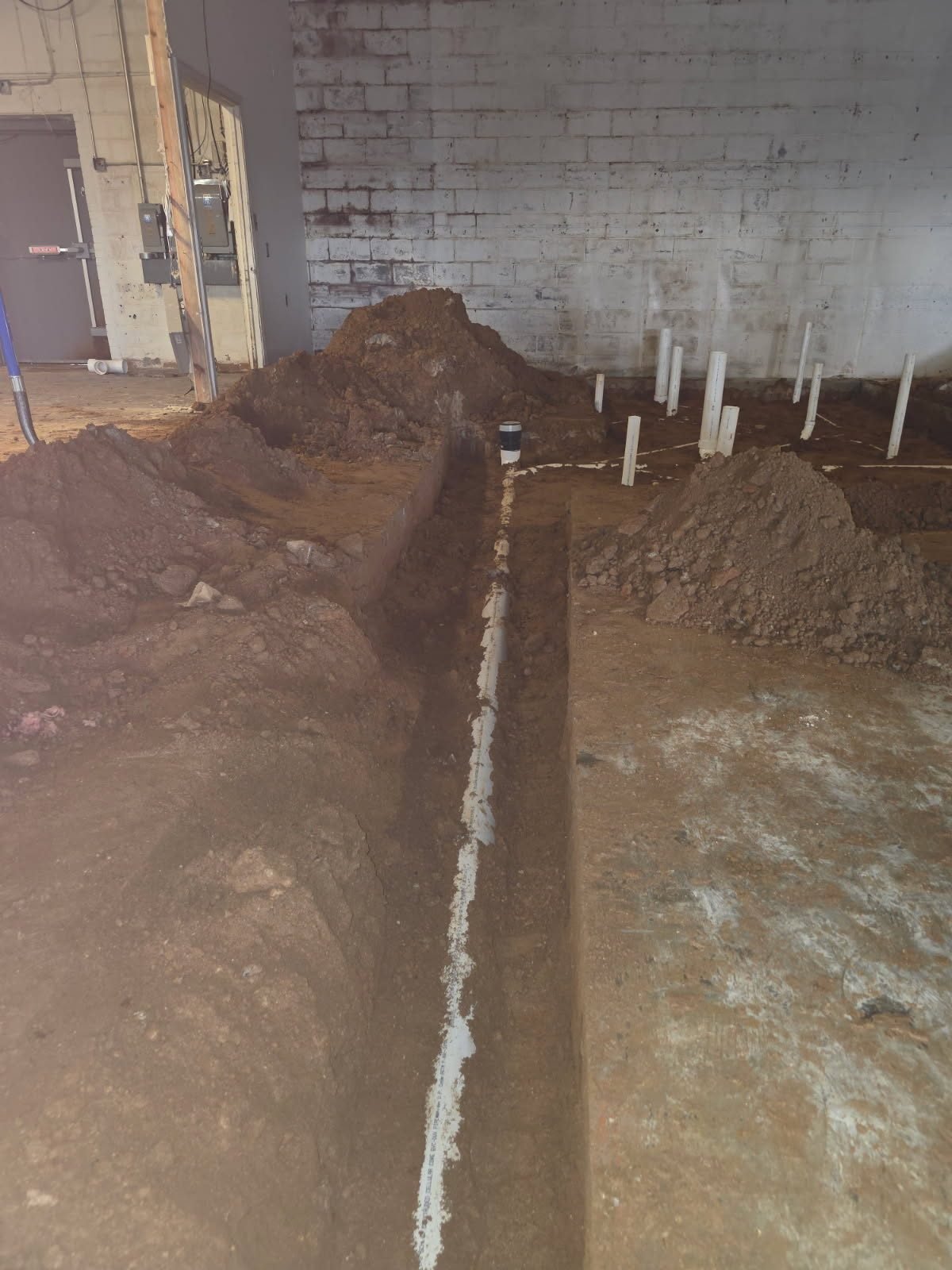 Interior trench leading to a row of PVC stub-up pipes near a concrete block wall.