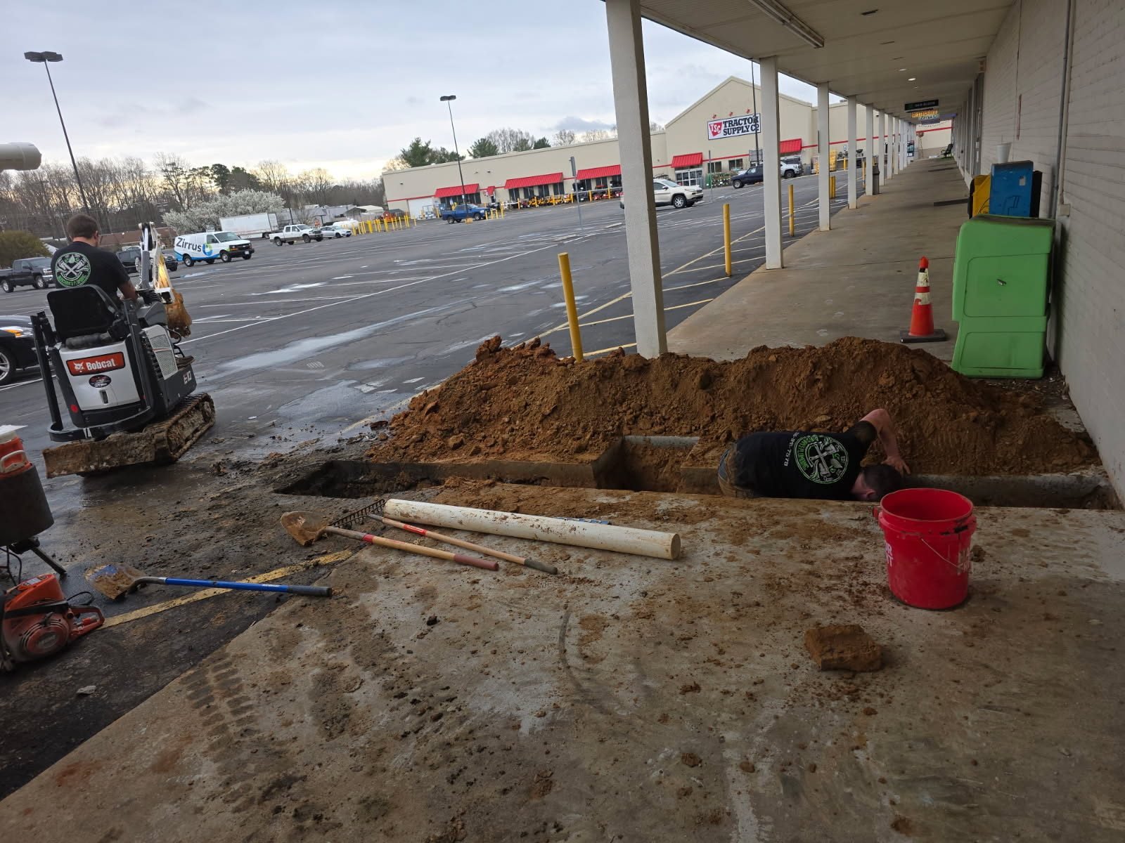 Worker kneeling in an exterior trench beneath a covered walkway, working on a PVC pipe.